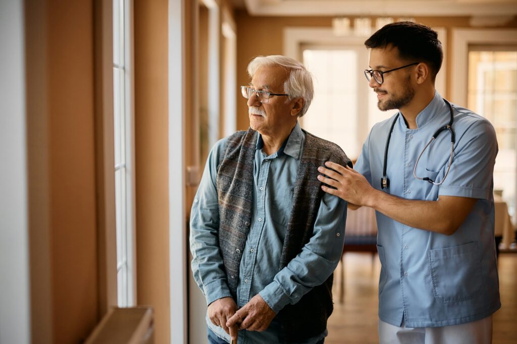 Male caregiver and senior man looking through the window at nursing home.
